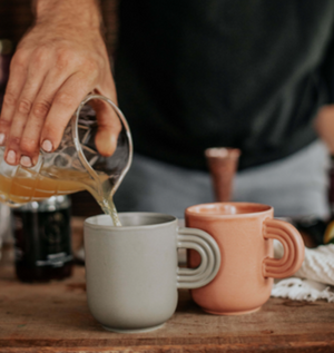 Close up of a hand pouring a Kvas Hot Ginger Toddy into a ceramic mug