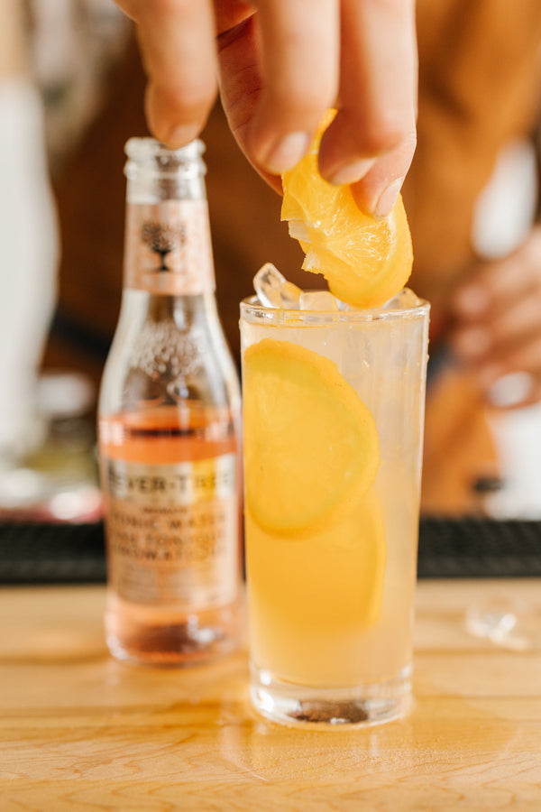 Close up of non-alcoholic Lavender Tonic cocktail with fresh orange slices and bottle of tonic water