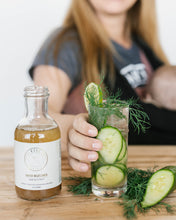 Load image into Gallery viewer, Kvas Honey Ginger Simple Syrup bottle next to craft cocktail garnished with cucumbers