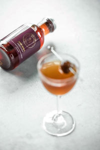 Closeup of Kvas Cherry Hibiscus Simple Syrup bottle on its side next to garnished cocktail